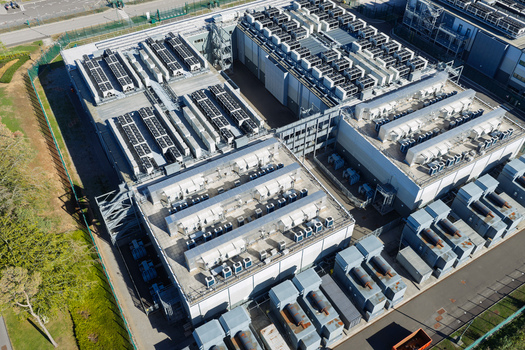Aerial view of a large data center facility with rows of industrial cooling units and ventilation systems on the rooftops surrounded by perimeter fencing in the United Kingdom.