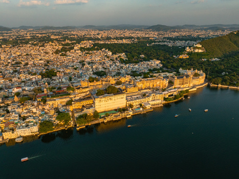 Aerial view of the historic City Palace complex on the banks of Lake Pichola with golden hour light illuminating the architecture and cityscape in Udaipur Rajasthan India.