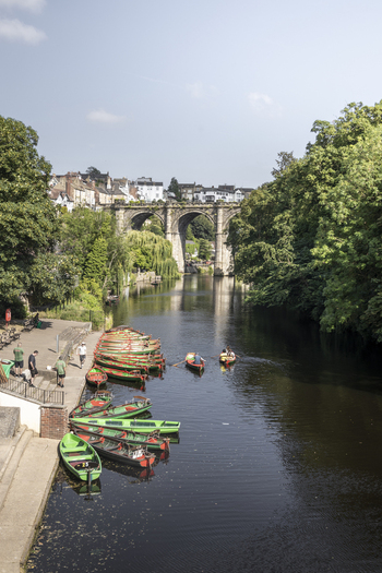 View of the historic Knaresborough Viaduct spanning the River Nidd with colorful rowing boats moored at the riverbank and people enjoying the water in Knaresborough England United Kingdom.
