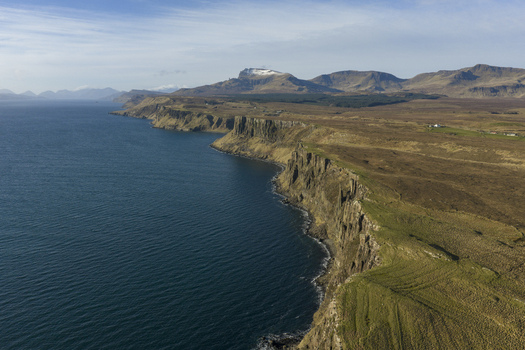 Aerial view of the rugged cliffs of the Trotternish peninsula and distant snow capped mountains under a clear sky in Isle of Skye Scotland.