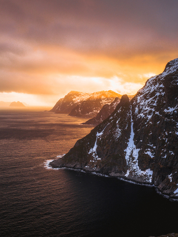 Aerial view of rugged snow dusted cliffs meet the dark rippling sea under a fiery sunset sky Lofoten Nordland Norway.