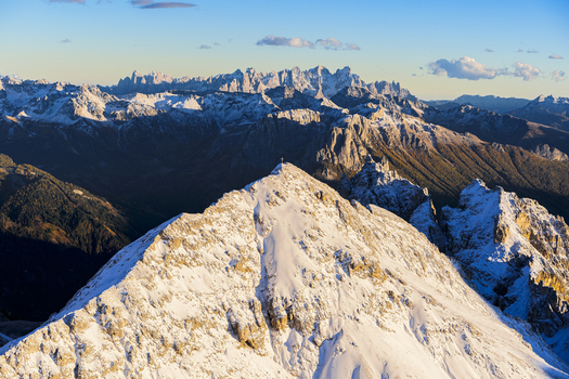 Aerial view of snow capped Catinaccio peak glowing under a warm sunset light contrasted by the deep shadows of the valleys below Trentino South Tyrol Italy.