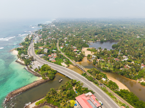 Aerial view of Devil s Rock with a coastal road bridge crossing a river surrounded by lush palm trees and turquoise ocean water in Ahangama Southern Province Sri Lanka. Aerial view of Devil s Rock with a coastal road bridge crossing a river surrounded by lush palm trees and turquoise ocean water in Ahangama Southern Province Sri Lanka.