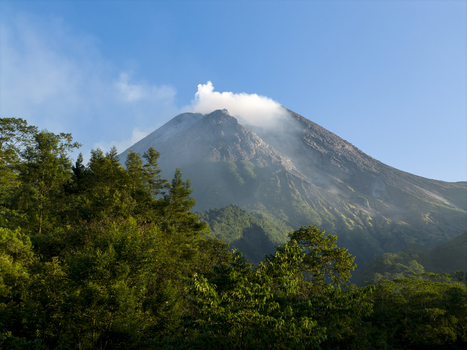 Aerial view of the majestic Mount Merapi volcano with smoke rising from its crater above lush green forests Kecamatan Pakem Daerah Istimewa Yogya Indonesia. Aerial view of the majestic Mount Merapi volcano with smoke rising from its crater above lush green forests Kecamatan Pakem Daerah Istimewa Yogya Indonesia.