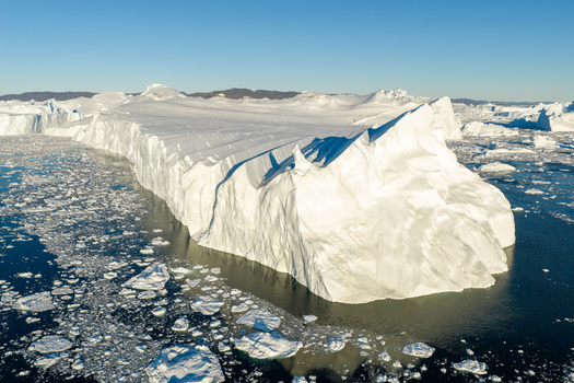 Aerial view of the massive Ilulissat glacier iceberg floating in dark blue arctic waters with scattered ice fragments under a clear sky in Greenland. Aerial view of the massive Ilulissat glacier iceberg floating in dark blue arctic waters with scattered ice fragments under a clear sky in Greenland.
