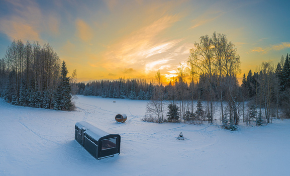 Aerial view of a small cabin and a barrel sauna in a snow covered forest landscape during a golden sunset in Latvia. Aerial view of a small cabin and a barrel sauna in a snow covered forest landscape during a golden sunset in Latvia.