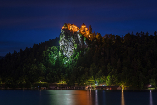 View of the illuminated Bled Castle perched on a steep cliff above the dark waters of Lake Bled at night in Bled Radovljica Slovenia. View of the illuminated Bled Castle perched on a steep cliff above the dark waters of Lake Bled at night in Bled Radovljica Slovenia.