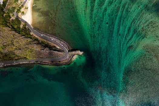 Aerial view of a winding coastal road with cars curving around a rocky outcrop next to vibrant turquoise coral reefs and clear ocean water in Mauritius. Aerial view of a winding coastal road with cars curving around a rocky outcrop next to vibrant turquoise coral reefs and clear ocean water in Mauritius.