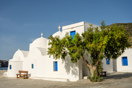 View of a traditional white washed Greek Orthodox church with blue doors and domes featuring a large tree in the foreground under a clear blue sky in Lefkada Greece.