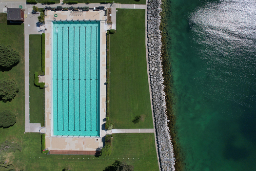 Aerial view of a rectangular outdoor swimming pool with turquoise water situated next to a rocky lakefront and lush green lawn in Morges Vaud Switzerland. Aerial view of a rectangular outdoor swimming pool with turquoise water situated next to a rocky lakefront and lush green lawn in Morges Vaud Switzerland.