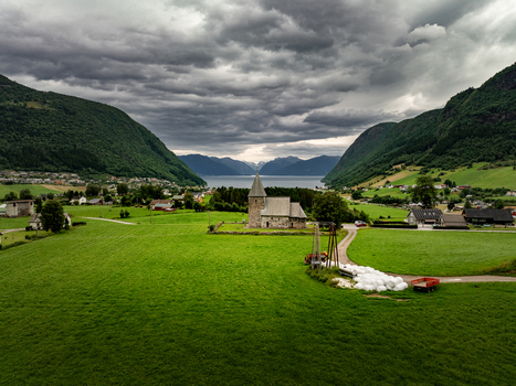 Aerial view of the historic stone Hove Church surrounded by lush green fields and rolling hills under a dramatic cloudy sky in Vik i Sogn Vestland Norway.
