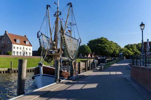 View of the historic Greetsiel harbor featuring a traditional shrimp cutter boat docked alongside brick houses and a green embankment under a clear blue sky in Krummhorn Lower Saxony Germany. View of the historic Greetsiel harbor featuring a traditional shrimp cutter boat docked alongside brick houses and a green embankment under a clear blue sky in Krummhorn Lower Saxony Germany.