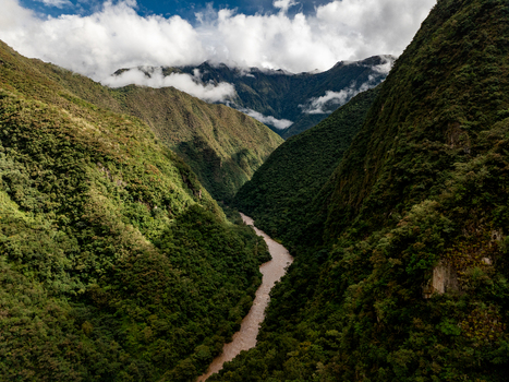 Aerial view of the Urubamba River winding through a deep lush green valley surrounded by steep Andean mountains under a cloudy sky in Aguas Calientes Cusco Peru. Aerial view of the Urubamba River winding through a deep lush green valley surrounded by steep Andean mountains under a cloudy sky in Aguas Calientes Cusco Peru.