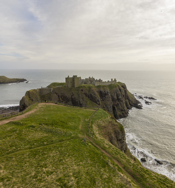 Aerial view of Dunnottar Castle medieval ruins perched on a rocky headland overlooking the North Sea with green grassy cliffs under a cloudy sky in Stonehaven Scotland United Kingdom. Aerial view of Dunnottar Castle medieval ruins perched on a rocky headland overlooking the North Sea with green grassy cliffs under a cloudy sky in Stonehaven Scotland United Kingdom.