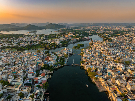 Aerial view of the historic city of Udaipur with its dense urban architecture shimmering lakes and distant hills under a warm golden sunset light in Udaipur Rajasthan India.