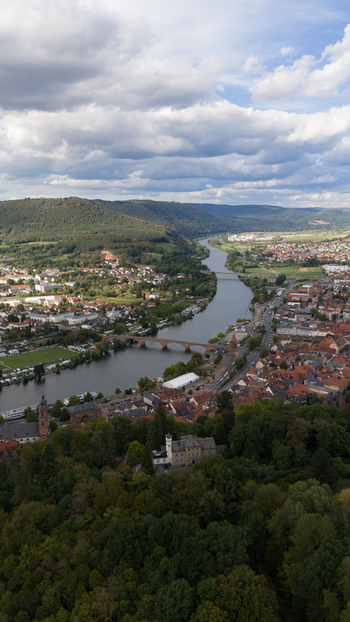 Aerial view of the Mainbrucke bridge spanning the Main river surrounded by historic town buildings lush green forests and rolling hills under a cloudy sky in Miltenberg Bayern Germany.