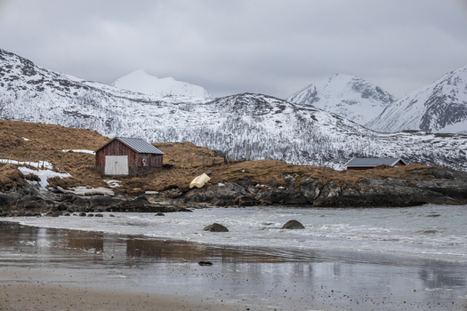 View of a traditional red wooden cabin on a rocky shoreline with snow capped mountains under an overcast sky in Kvaloya Troms Norway. View of a traditional red wooden cabin on a rocky shoreline with snow capped mountains under an overcast sky in Kvaloya Troms Norway.