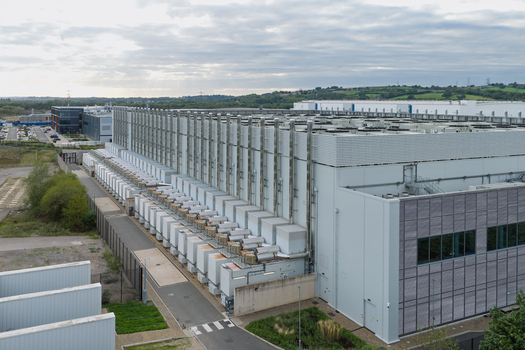 Aerial view of a large data center facility featuring rows of external cooling units modern metal cladding and surrounding green landscape United Kingdom.