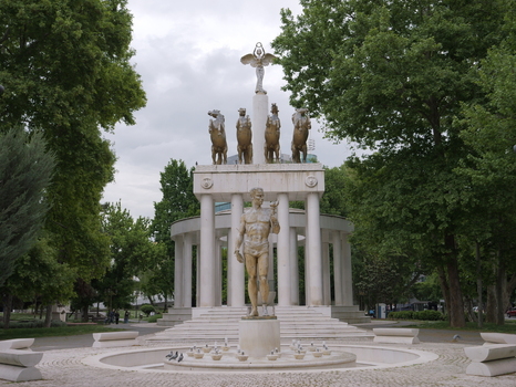 View of the Warrior on a Horse monument featuring a central male statue and a colonnade with a quadriga sculpture surrounded by lush green trees in Skopje Greater Skopje North Macedonia.
