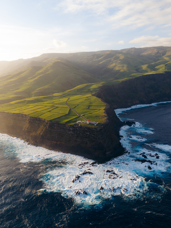 Aerial view of Farol de Albarnaz lighthouse on a steep cliff edge with green fields and mountains under golden sunlight in Ponta Delgada Acores Portugal. Aerial view of Farol de Albarnaz lighthouse on a steep cliff edge with green fields and mountains under golden sunlight in Ponta Delgada Acores Portugal.