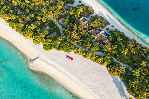 Aerial view of a tropical island resort with white sand beaches turquoise waters lush palm trees and villas with red umbrellas on the sand near Kudahuvadhoo Dhaalu Atoll Maldives. Aerial view of a tropical island resort with white sand beaches turquoise waters lush palm trees and villas with red umbrellas on the sand near Kudahuvadhoo Dhaalu Atoll Maldives.