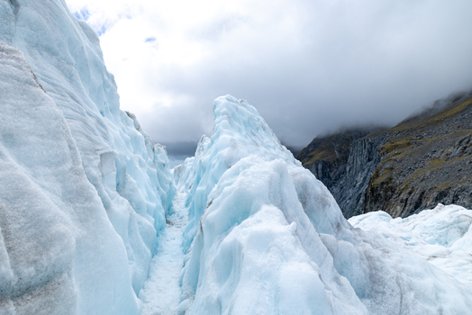 View of the icy blue glacier contrasting with the rugged rocky terrain under a cloudy sky creating a serene yet powerful landscape Franz Josef Glacier West Coast Region New Zealand. View of the icy blue glacier contrasting with the rugged rocky terrain under a cloudy sky creating a serene yet powerful landscape Franz Josef Glacier West Coast Region New Zealand.