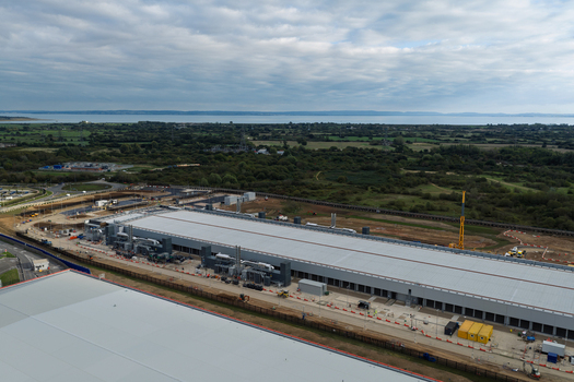 Aerial view of a large data center facility with a long grey roof industrial equipment and surrounding green fields and trees in the United Kingdom.