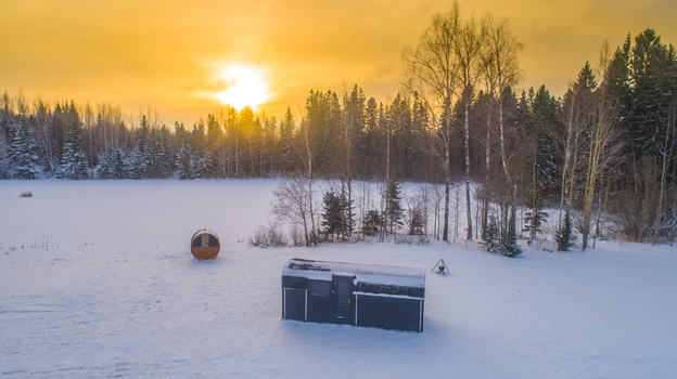 Aerial view of a small dark cabin and a barrel sauna in a snow covered landscape with a forest backdrop during a golden sunset in Latvia. Aerial view of a small dark cabin and a barrel sauna in a snow covered landscape with a forest backdrop during a golden sunset in Latvia.