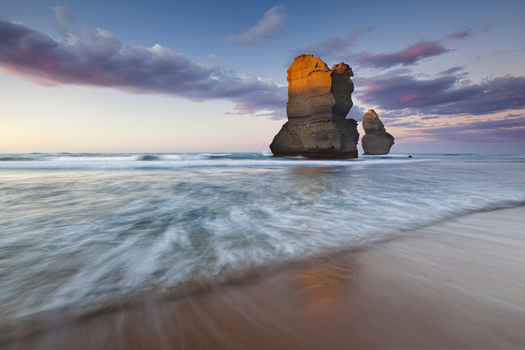 View of the Twelve Apostles limestone stacks standing in the ocean with waves crashing on the sandy beach under a sunset sky in Twelve Apostles Marine National Park Australia. View of the Twelve Apostles limestone stacks standing in the ocean with waves crashing on the sandy beach under a sunset sky in Twelve Apostles Marine National Park Australia.