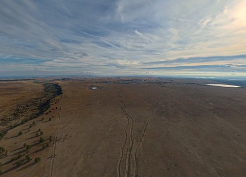 Aerial view of the vast dry prairie landscape with visible tire tracks and a small pond under an expansive cloudy sky in Golden Colorado United States.