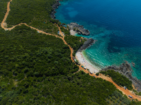 Aerial view of Aquarium Beach featuring turquoise water rocky cliffs and a winding dirt road through lush green hills in Jala Vlore County Albania. Aerial view of Aquarium Beach featuring turquoise water rocky cliffs and a winding dirt road through lush green hills in Jala Vlore County Albania.