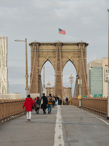 View of the Brooklyn Bridge with pedestrians walking along the wooden walkway stone arches and the American flag flying under an overcast sky in Kings County New York United States.