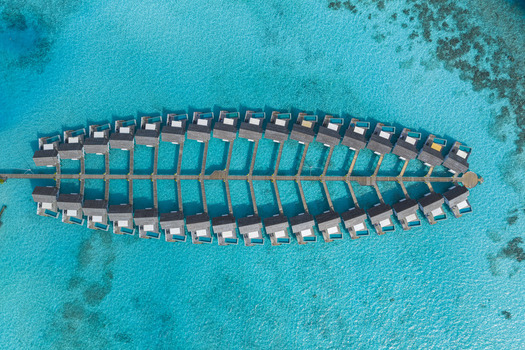Aerial view of luxury overwater bungalows arranged in a symmetrical pattern over a turquoise lagoon with visible coral reefs Maldives.