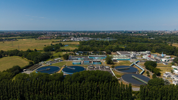 Aerial view of a wastewater treatment plant with circular sedimentation tanks surrounded by lush green trees and fields with the city skyline of Milan in the distance Milan Lombardy Italy. Aerial view of a wastewater treatment plant with circular sedimentation tanks surrounded by lush green trees and fields with the city skyline of Milan in the distance Milan Lombardy Italy.