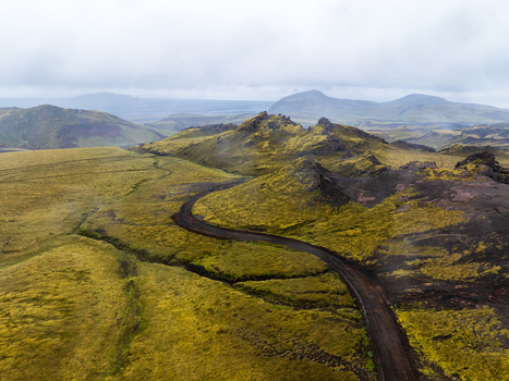 Aerial view of a winding dirt road cutting through a vast moss covered volcanic landscape with distant mountains under a cloudy sky in Iceland. Aerial view of a winding dirt road cutting through a vast moss covered volcanic landscape with distant mountains under a cloudy sky in Iceland.