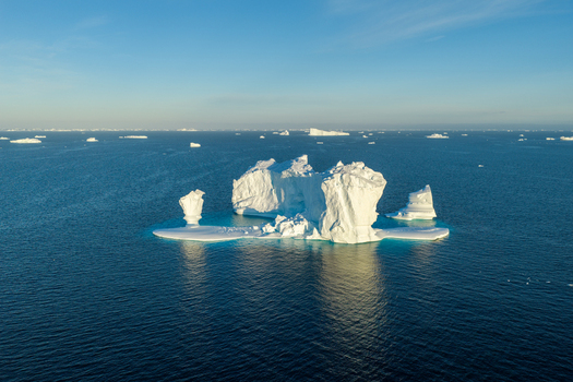 Aerial view of an arch iceberg floating in the deep blue ocean surrounded by smaller icebergs under a clear sky in Greenland. Aerial view of an arch iceberg floating in the deep blue ocean surrounded by smaller icebergs under a clear sky in Greenland.