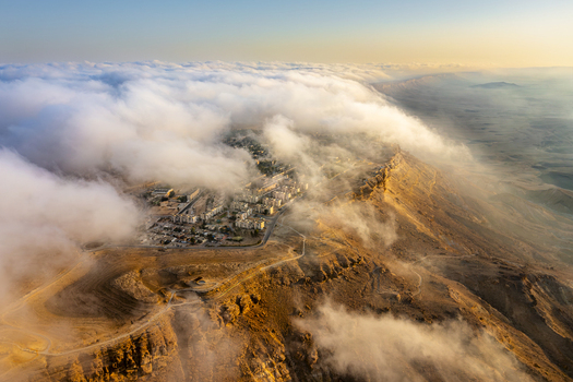 Aerial view of Mitzpe Ramon crater partially covered by low hanging clouds at sunset with desert cliffs and town buildings in Mitzpe Ramon South District Israel. Aerial view of Mitzpe Ramon crater partially covered by low hanging clouds at sunset with desert cliffs and town buildings in Mitzpe Ramon South District Israel.