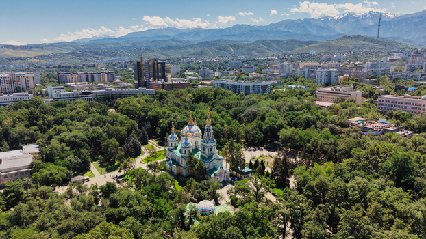 Aerial view of the colorful Ascension Cathedral surrounded by lush green trees in Panfilov Park with the city skyline and mountains in the background in Almaty Almaty Region Kazakhstan.