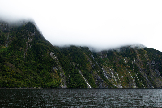 View of dramatic mist shrouded mountains rise majestically from the dark still waters waterfalls cascade down the verdant slopes Milford Sound Southland Region New Zealand.