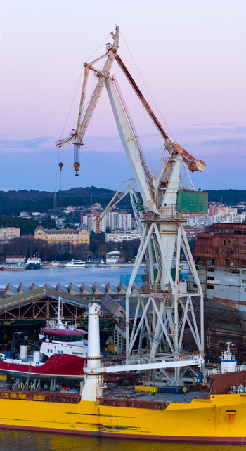 Aerial view of the Uljanik Shipyard featuring a large industrial crane a yellow cargo ship and a red tugboat docked in the harbor of Pula Croatia.