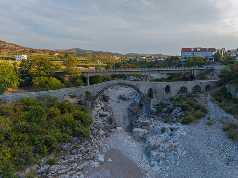 Aerial view of the historic Mesi Bridge spanning a dry rocky riverbed with lush vegetation and a modern bridge in the background in Boks Qarku i Shkodres Albania.