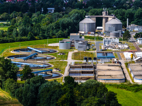 Aerial view of a wastewater treatment plant with circular sedimentation tanks and rectangular basins surrounded by green fields and trees in Cuxhaven Niedersachsen Germany.