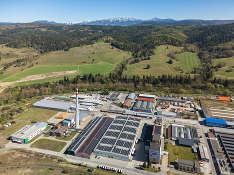 Aerial view of an industrial facility with solar panels on rooftops and a tall chimney set against green hills and distant snow capped mountains in Nizna Zilina Region Slovakia. Aerial view of an industrial facility with solar panels on rooftops and a tall chimney set against green hills and distant snow capped mountains in Nizna Zilina Region Slovakia.