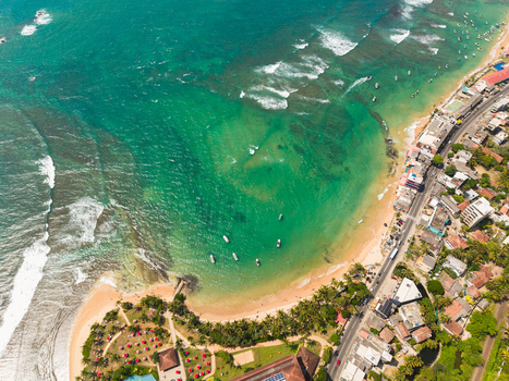 Aerial view of the turquoise coastline with sandy beaches palm trees and small boats anchored in the clear water in Hikkaduwa Southern Province Sri Lanka. Aerial view of the turquoise coastline with sandy beaches palm trees and small boats anchored in the clear water in Hikkaduwa Southern Province Sri Lanka.