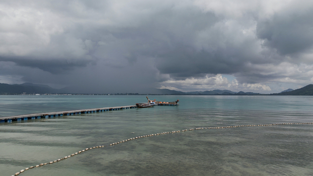 Aerial view of a wooden pier extending into calm turquoise water with traditional long tail boats moored at the end under a dramatic stormy sky near the tropical coast of Thailand. Aerial view of a wooden pier extending into calm turquoise water with traditional long tail boats moored at the end under a dramatic stormy sky near the tropical coast of Thailand.