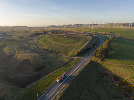Aerial view of a truck driving on a highway through rolling green hills with a large wind farm in the background under a clear sky during golden hour. Aerial view of a truck driving on a highway through rolling green hills with a large wind farm in the background under a clear sky during golden hour.