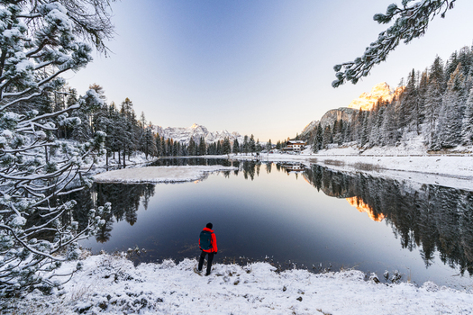 View of a solitary figure in a vibrant red jacket stands on the snowy shores of a serene lake mirroring the snow dusted evergreens and distant majestic mountains Misurina Veneto Italy. View of a solitary figure in a vibrant red jacket stands on the snowy shores of a serene lake mirroring the snow dusted evergreens and distant majestic mountains Misurina Veneto Italy.