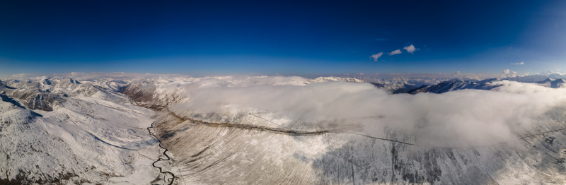 Aerial view of Babusar Pass winding through snow capped mountain peaks under a clear blue sky with rolling clouds in Babusar KPK Pakistan.