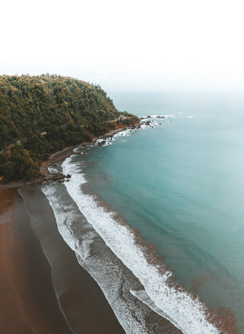 Aerial view of a pristine coastline with turquoise ocean waves crashing against a sandy beach and a lush green forested cliffside in Costa Rica.