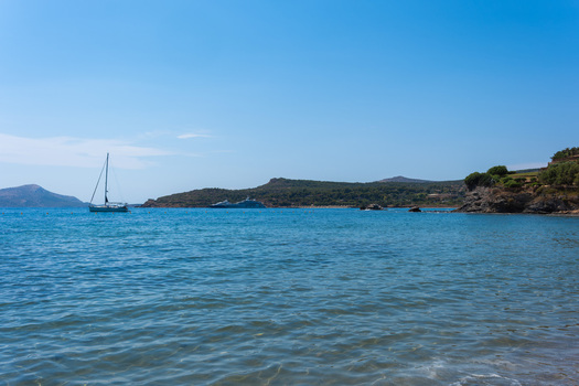View of a sailboat and yacht on the calm blue waters of the Aegean Sea near the rocky coastline of Cape Sounio in Sounion Greece.
